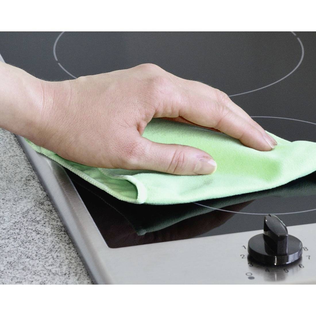 A hand wipes a clean, black ceramic hob with a green cloth, with a silver control knob. A kitchen worktop is beside it.