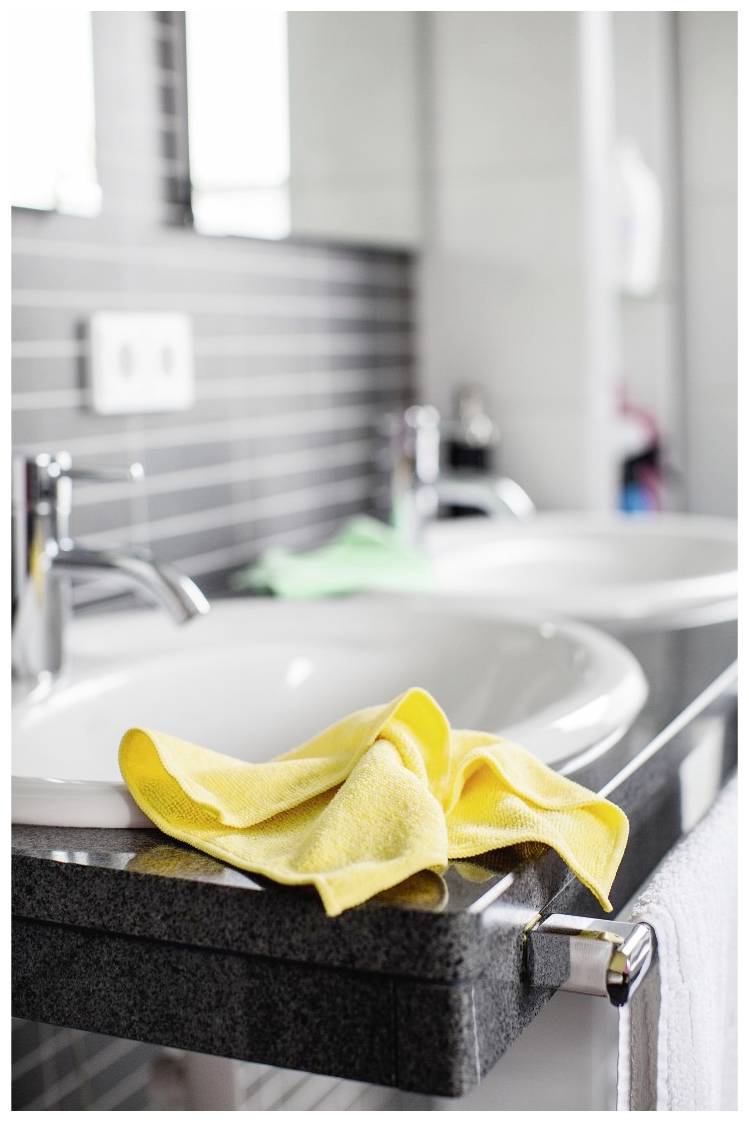 Yellow cleaning cloth on a bathroom worktop between two washbasins with taps, behind which is a tiled wall and mirror.