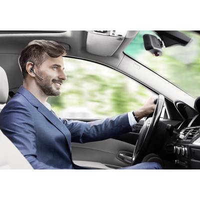 A man in a suit is driving a car and wearing a Bluetooth headset, looking straight ahead with a smile.