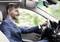 A man in a suit is driving a car and wearing a Bluetooth headset, looking straight ahead with a smile.
