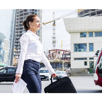 A woman in business attire is crossing the street in an urban area with construction sites in the background, carrying a bag and some papers.