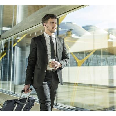 A man in a suit is pulling a wheeled suitcase and holding a coffee cup while walking through a modern airport terminal.