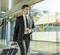 A man in a suit is pulling a wheeled suitcase and holding a coffee cup while walking through a modern airport terminal.