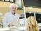 An elderly man in a white lab coat and hairnet serves a customer in a delicatessen shop.