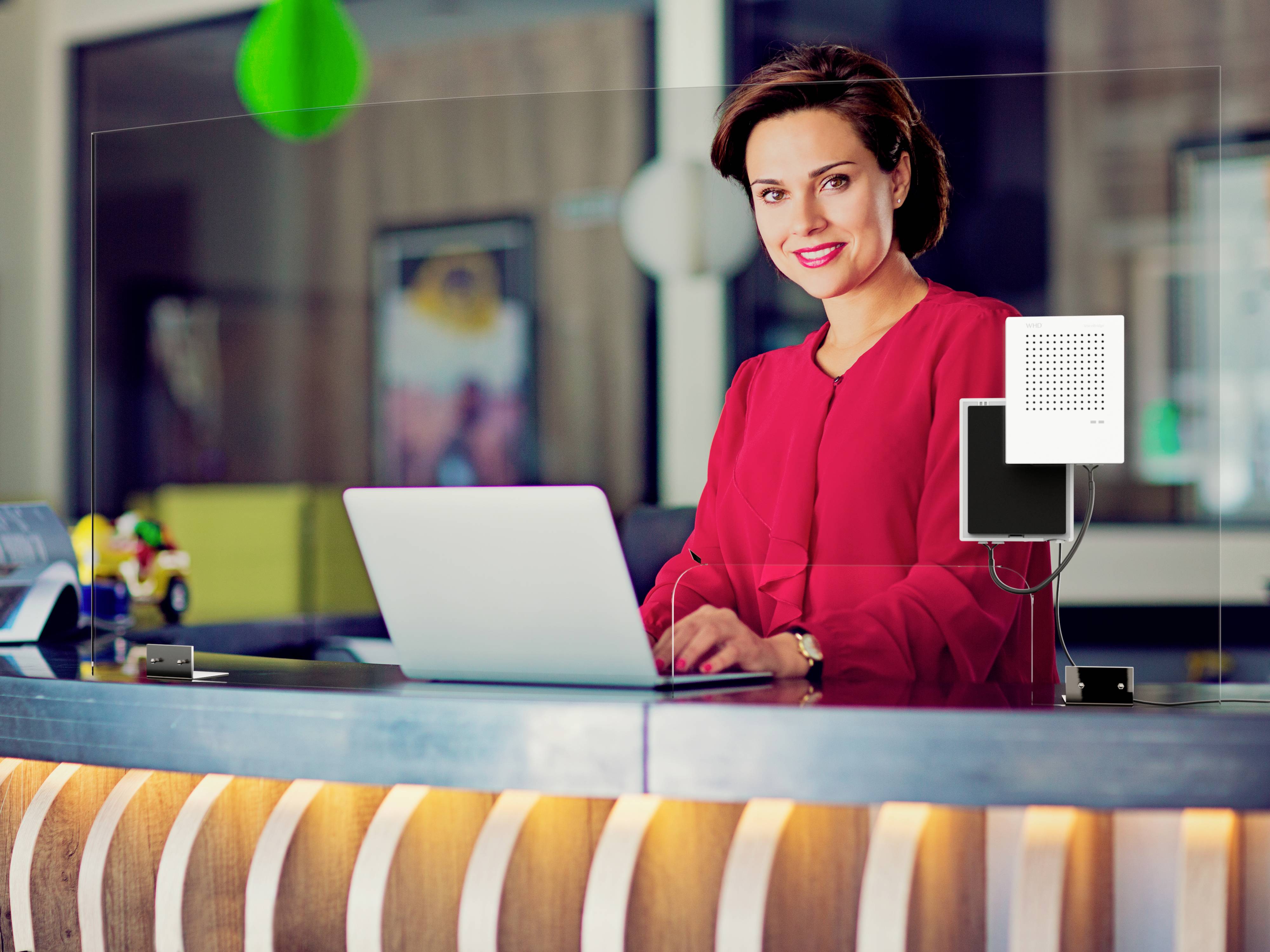 A woman in a red top is working on a laptop at a reception desk. Decorations and a transparent protective glass are visible in the background.