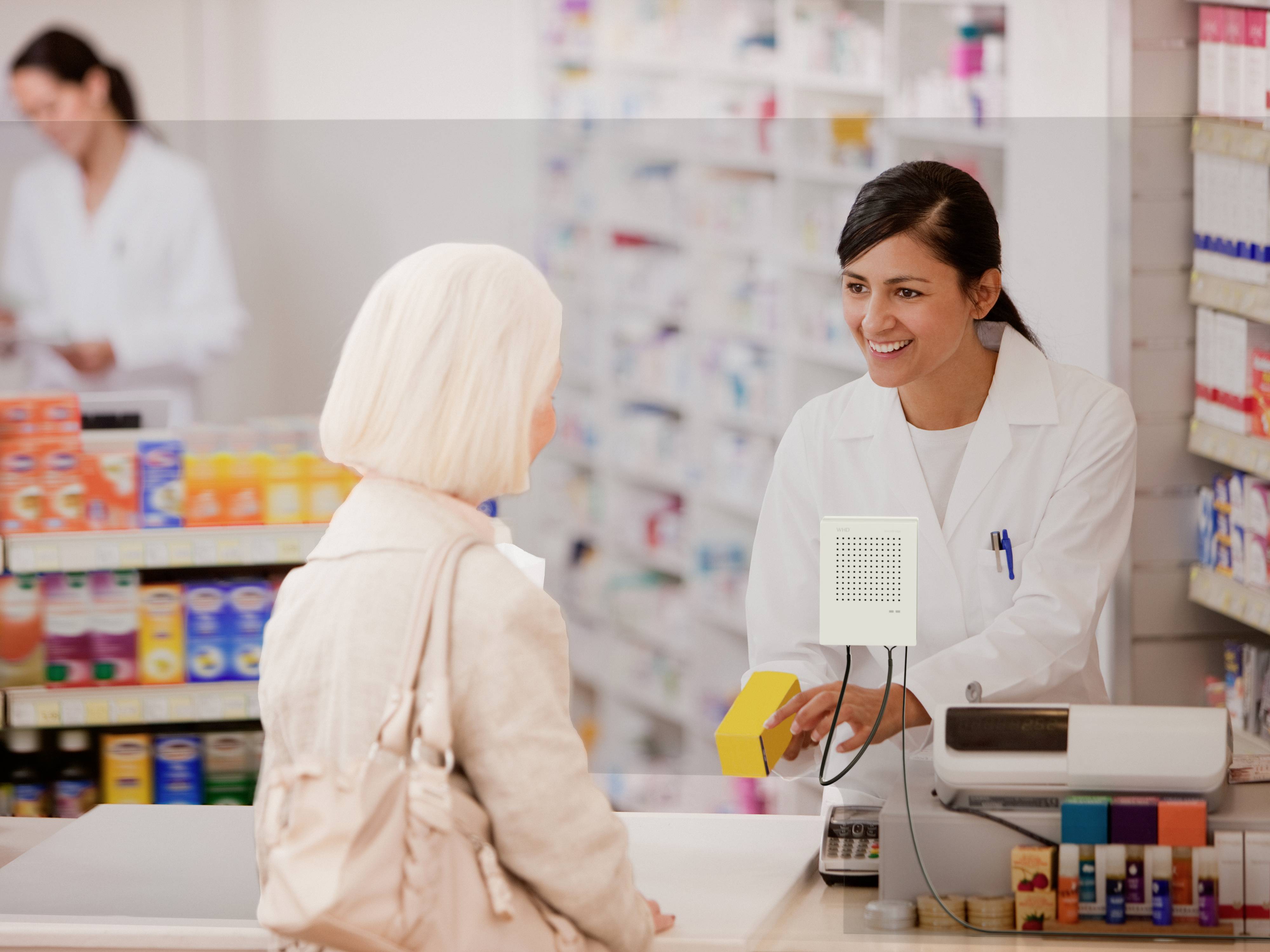 A female pharmacist hands a yellow medication to an elderly female customer across the counter. Shelves filled with products are visible in the background.