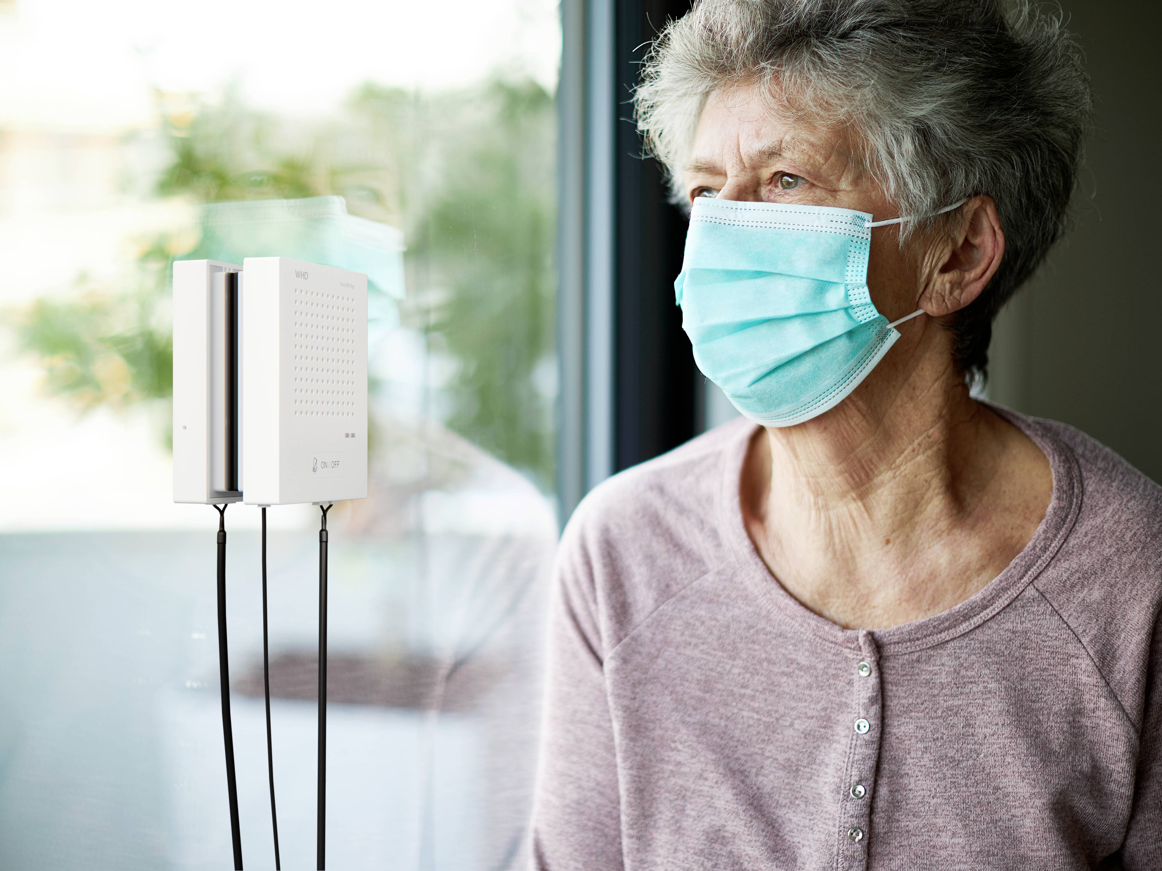 An elderly woman wearing a mask looks out of a window, next to a small device with cables, possibly a medical or environmental monitoring device.