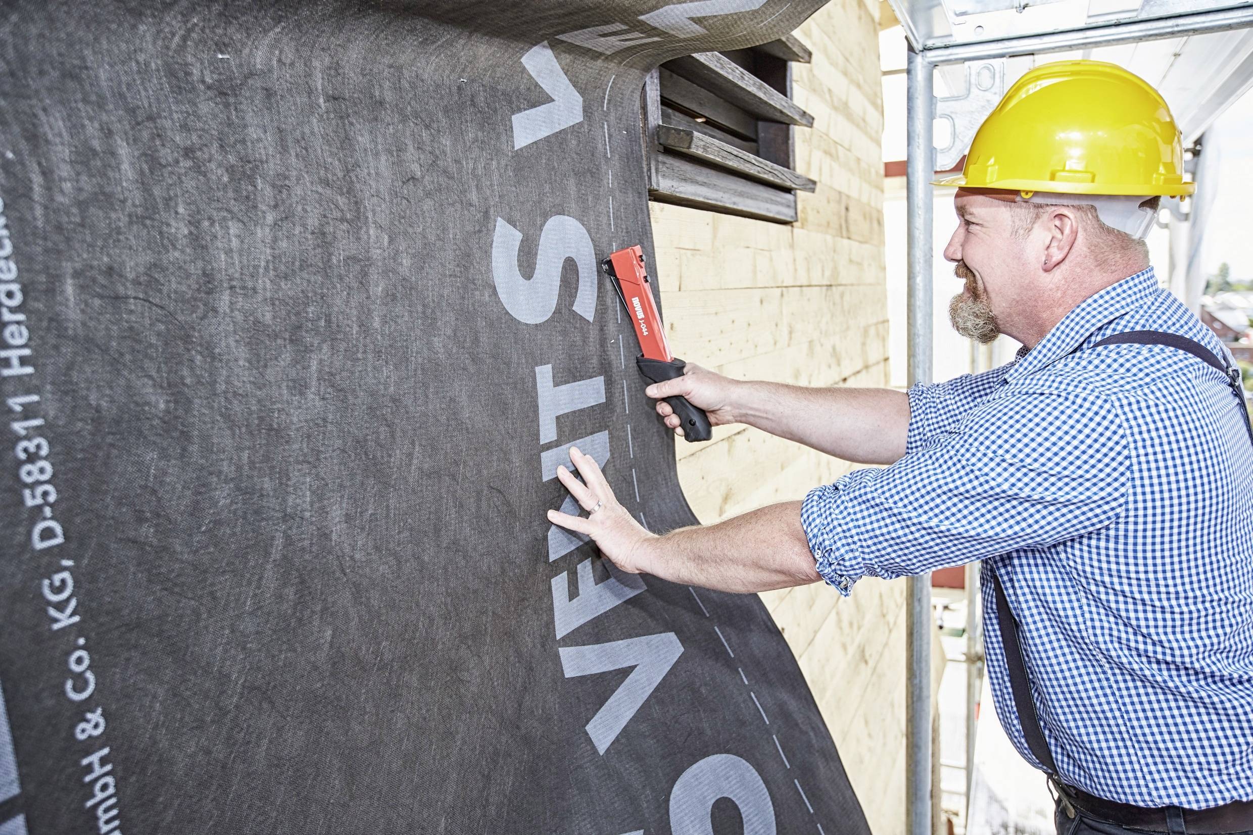 A construction worker is attaching a black insulating membrane to the exterior wall of a building. He is wearing a yellow hard hat.