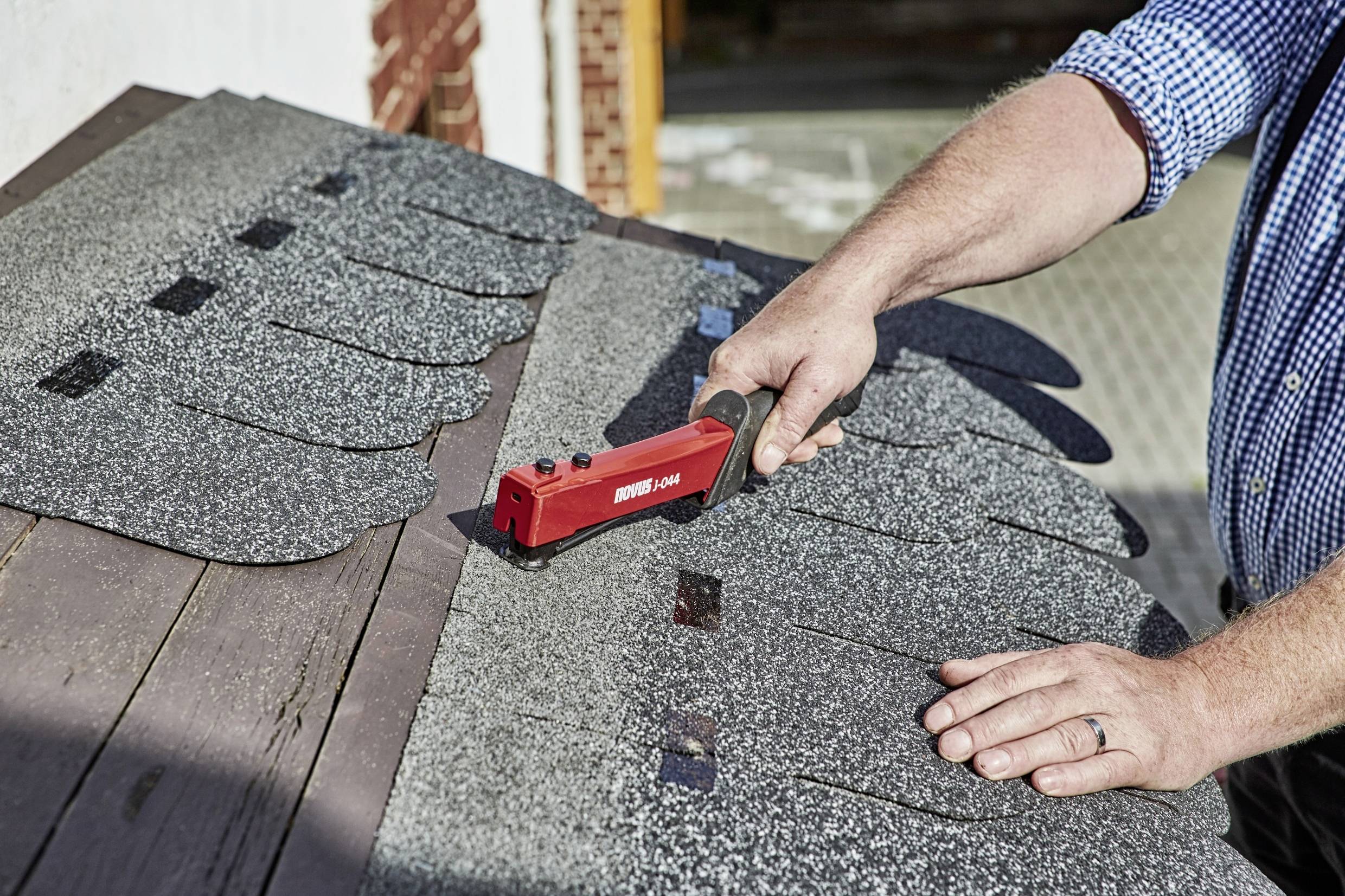 A person is using a red tool to work on roof tiles on a wooden roof. A paved path is visible in the background.