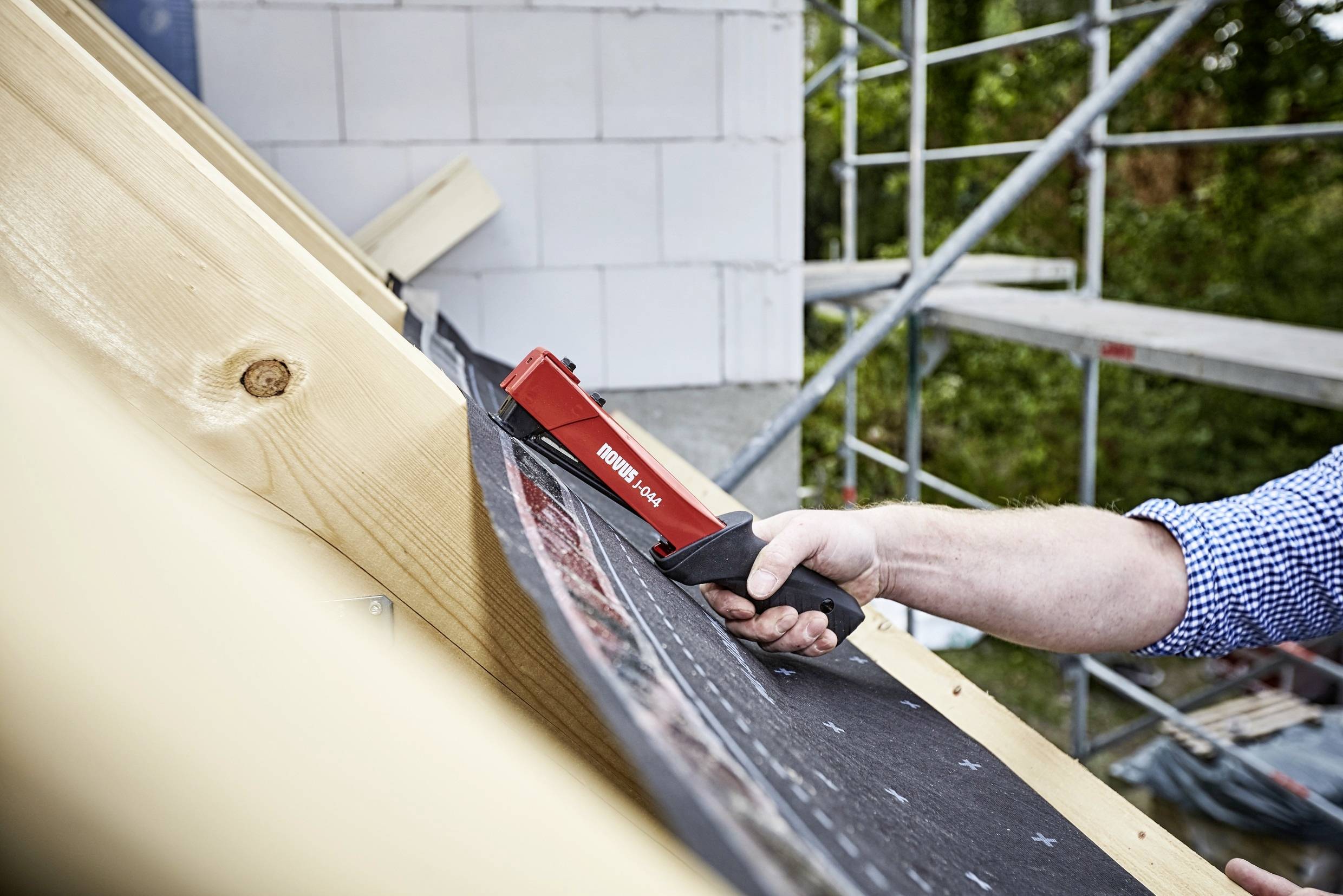 A person is securing roof membrane with a red hand tool on a wooden structure. Scaffolding is visible in the background.
