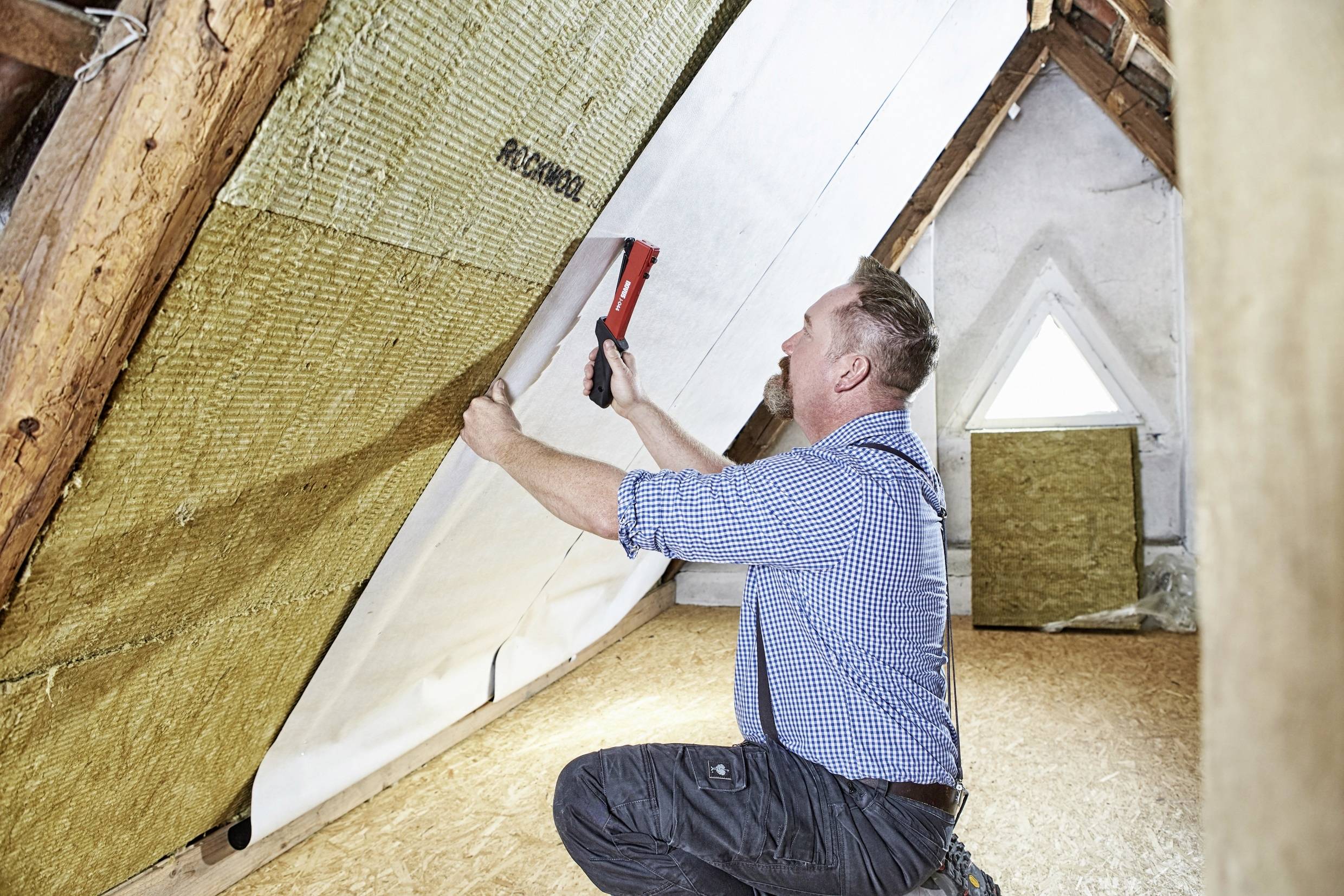 A man is insulating an attic with insulation material. He is using a staple gun to secure the material. A small window is visible in the background.