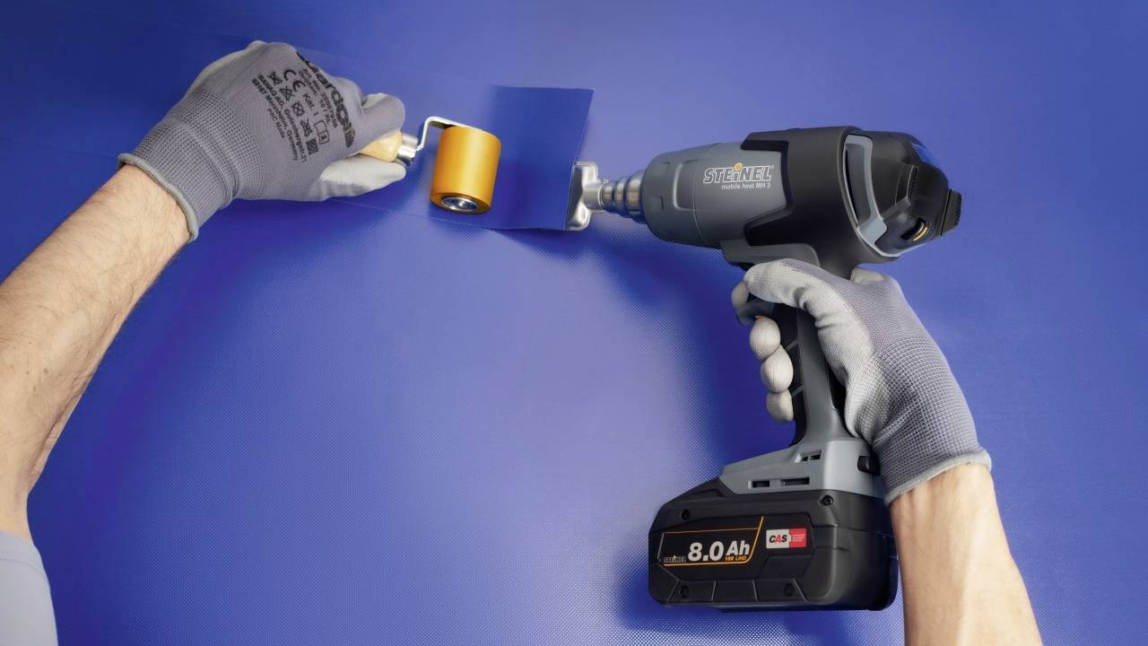 'A person is using an electric device to attach a blue wallpaper to the wall. On the left, a hand with a roller tool is visible.'