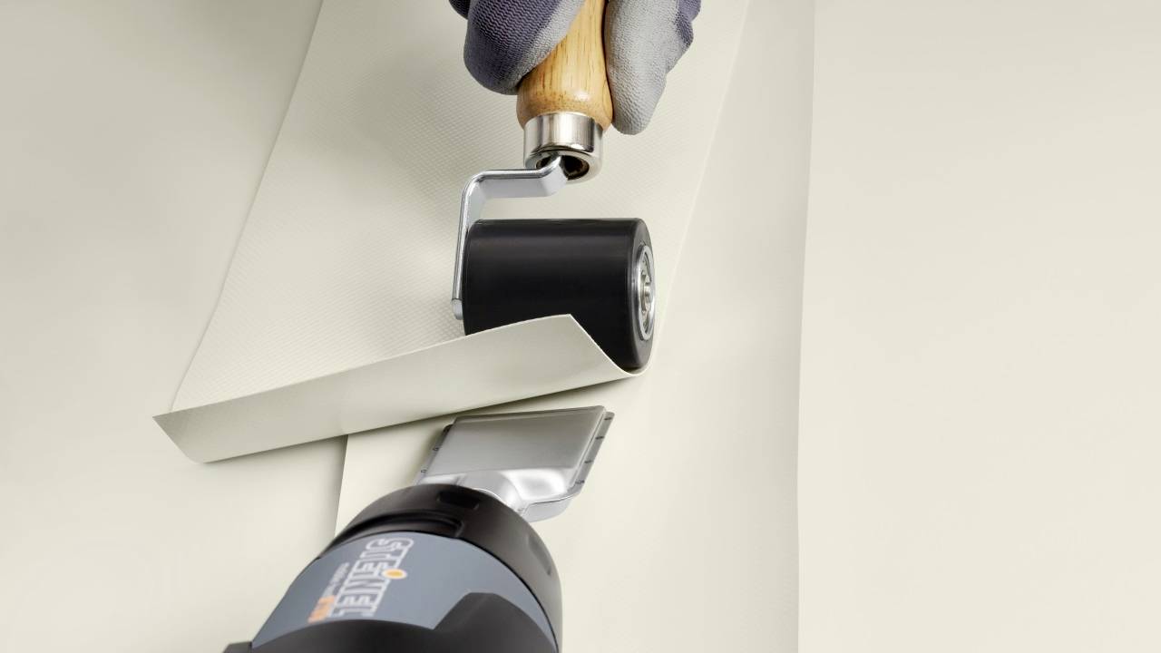 'Close-up of hands using a roller tool to press a wallpaper edge against the wall; an edge roller and wallpaper smoother are in use.'