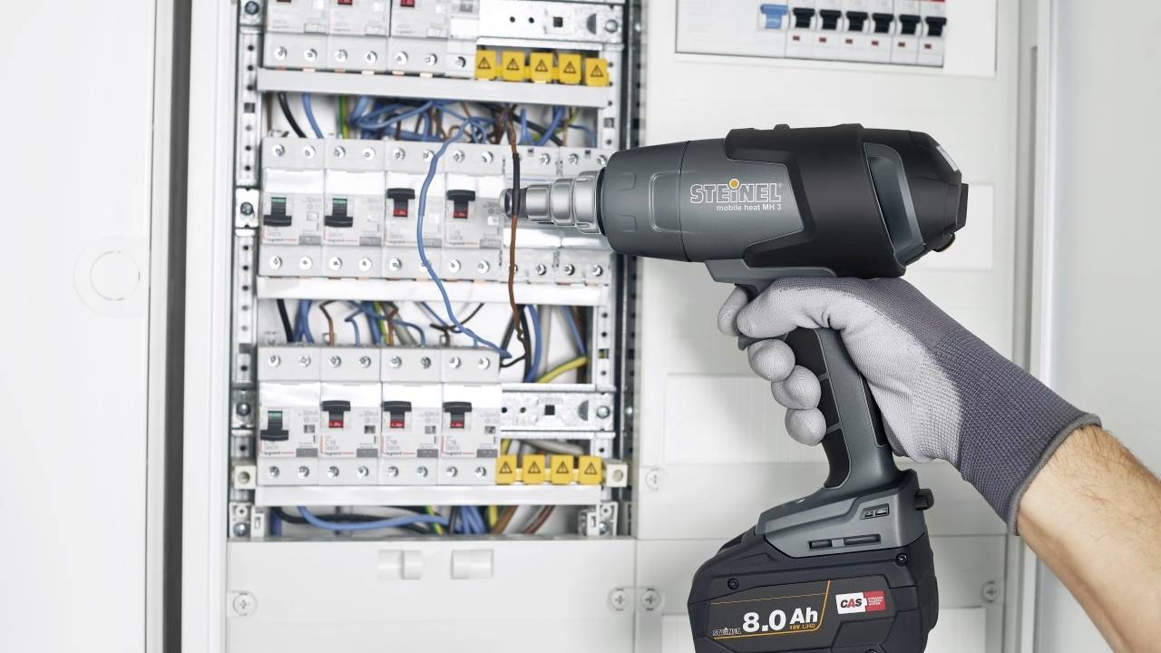 A person wearing gloves is holding a cordless drill while working on an open fuse box.