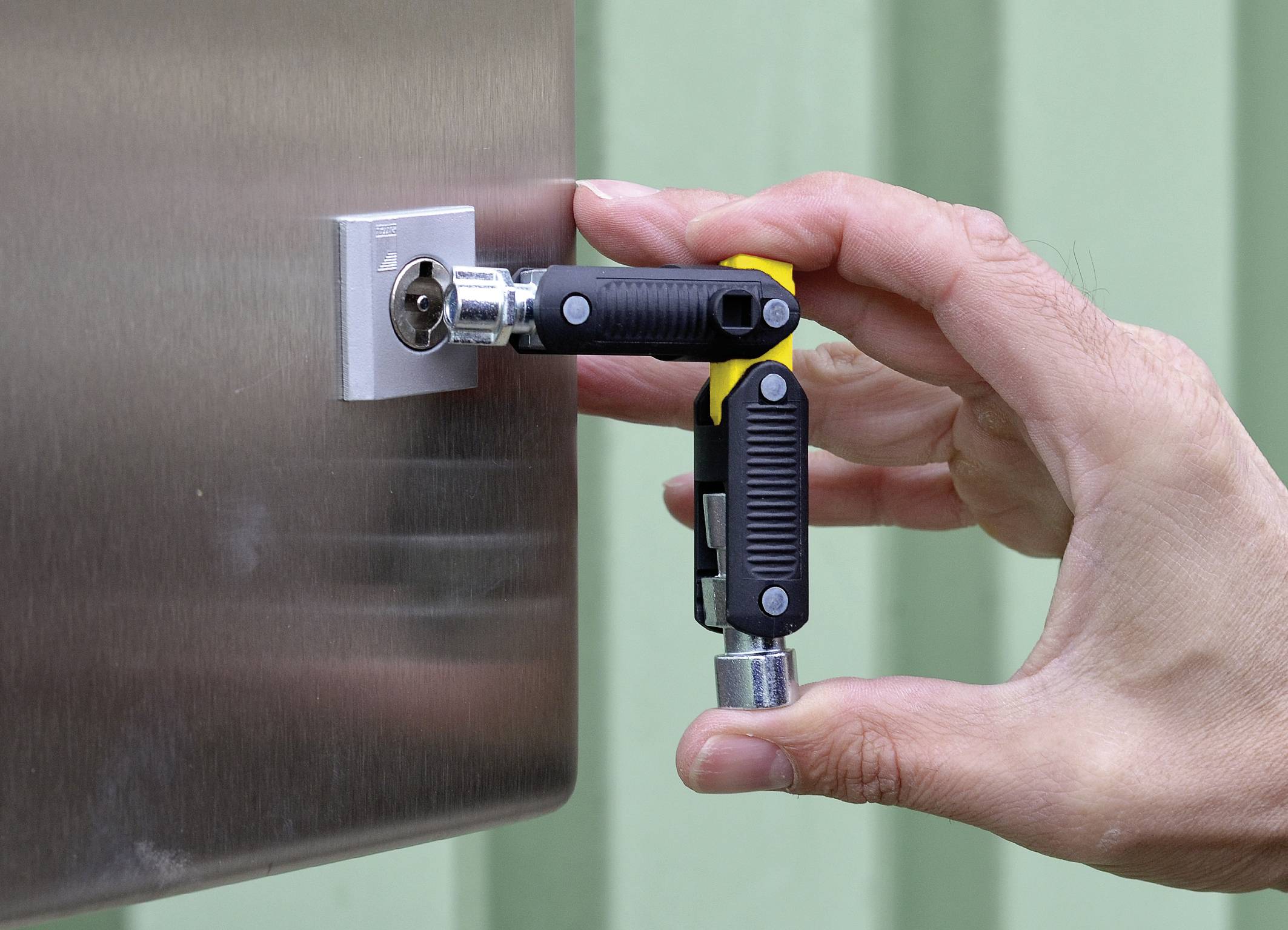A hand is using a tool to manipulate a lock on a metallic surface. The background shows a green wall.