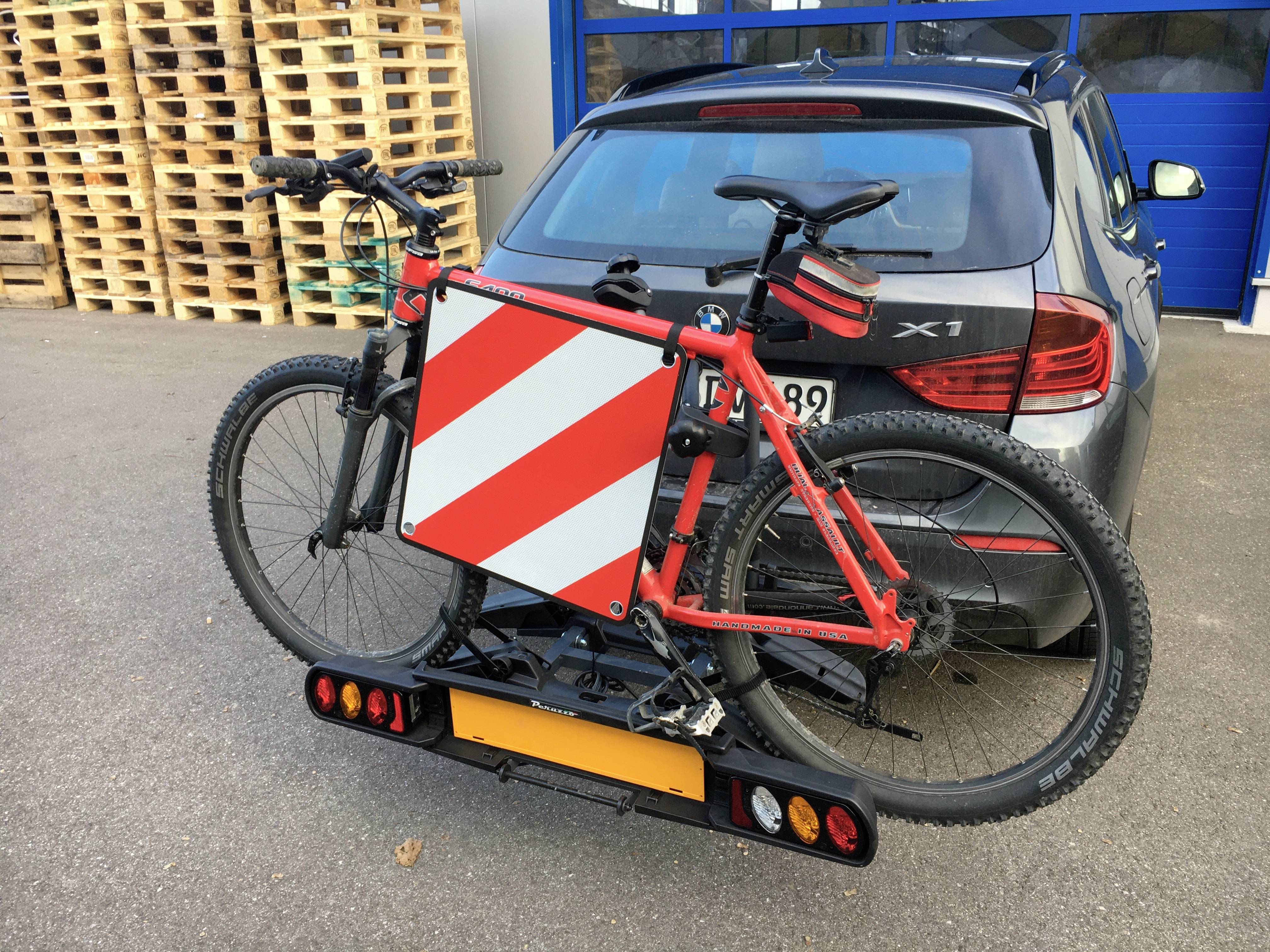 Grey car with a bicycle rack and a red bicycle mounted. Warning sign attached to the rack, parked in front of a building with stacks of pallets.