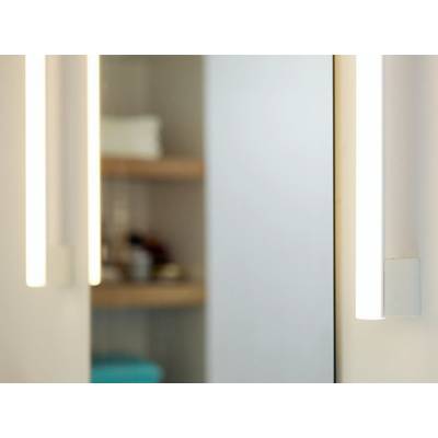 Illuminated mirror with two vertical lights on the sides. In the background, blurry shelves with various objects are visible.