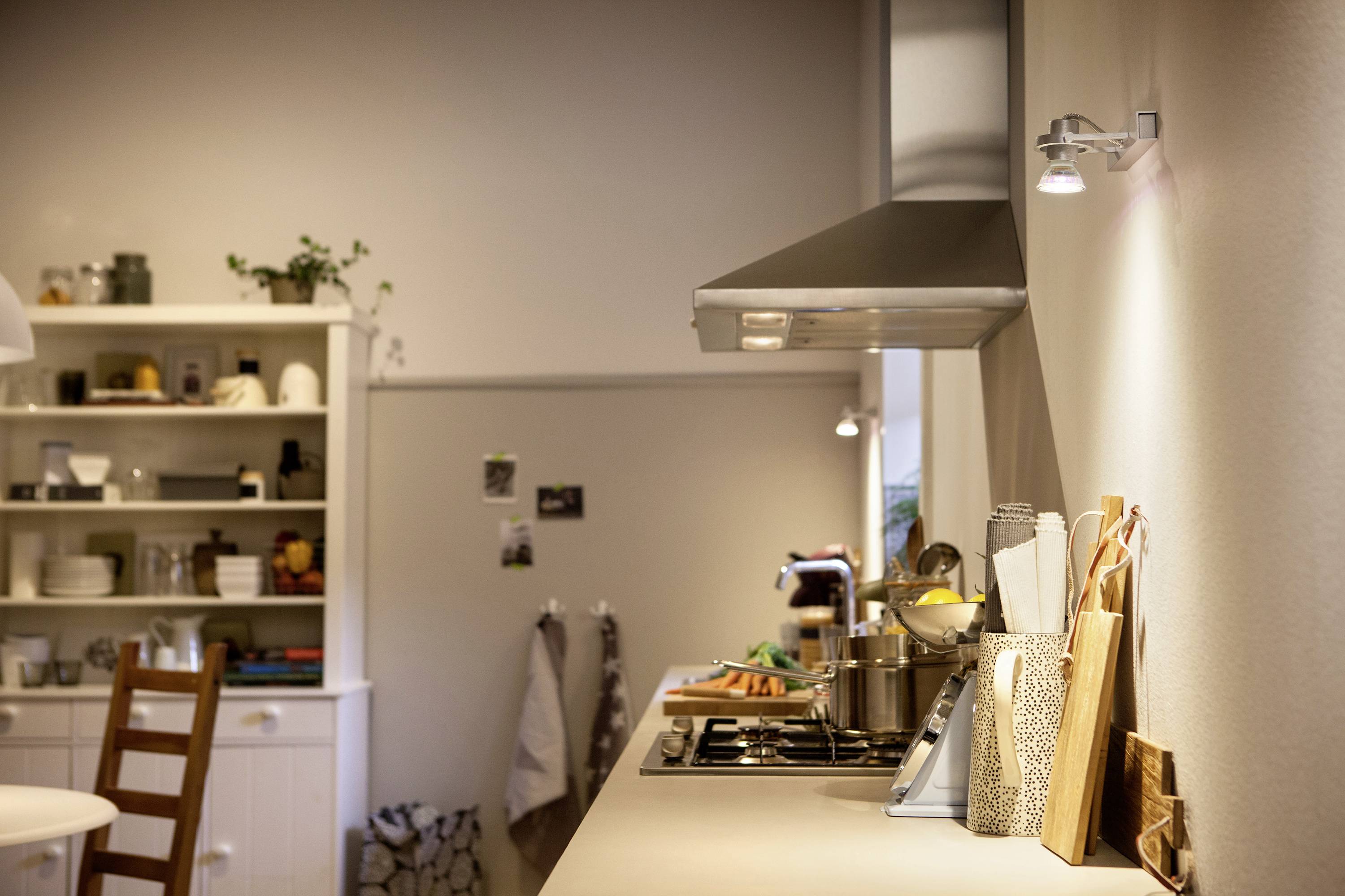 A cosy kitchen with a worktop laden with kitchen utensils. In the background, a shelf with decorative items and a dining table.