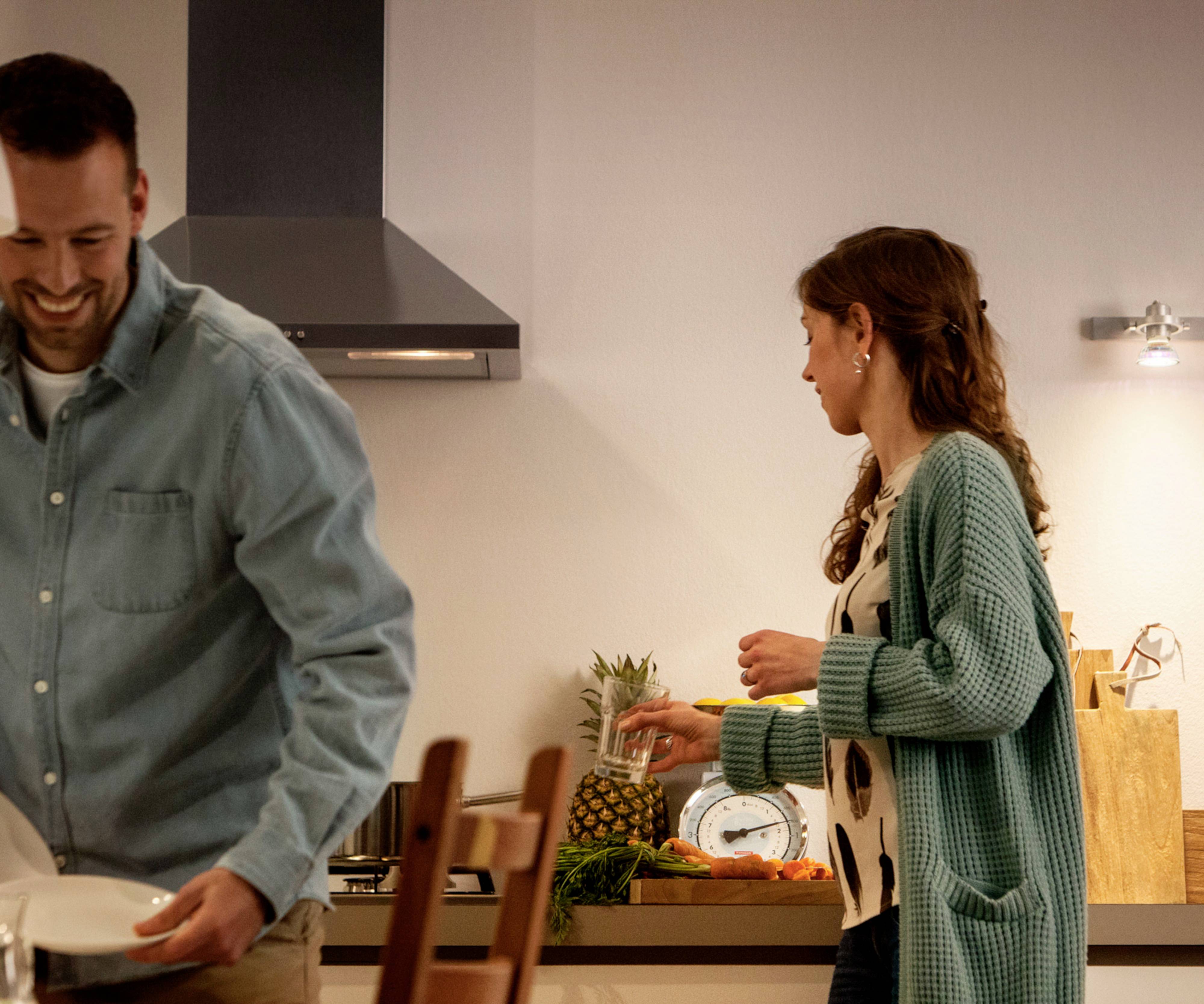 A man and a woman in a kitchen, the man smiling and holding a plate, the woman standing at the worktop next to fruit and a scales.