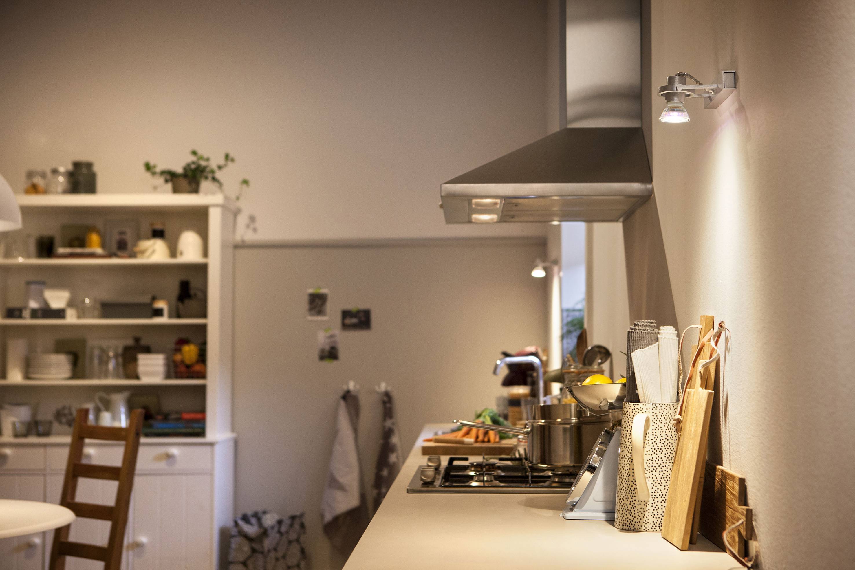 A modern kitchen with bright interior. On the left, a shelf with crockery and plants, on the right a worktop with cooking utensils and vegetables.