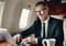 A man in a suit is smiling while working on a laptop inside an aircraft. A coffee cup sits on the table beside him.