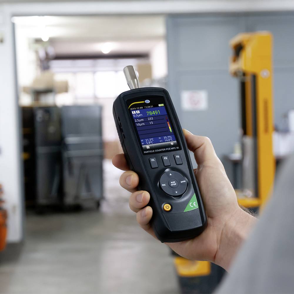 A hand holds a portable measuring device in a warehouse, displaying environmental values on a digital screen. Cardboard boxes and shelves are visible in the background.