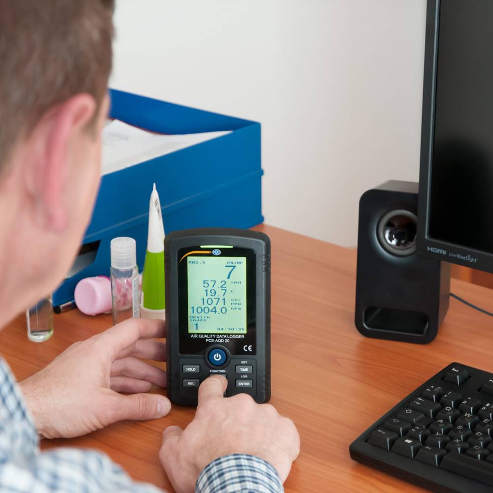 A person is using a portable air quality measuring device on a desk next to a computer screen and office supplies.