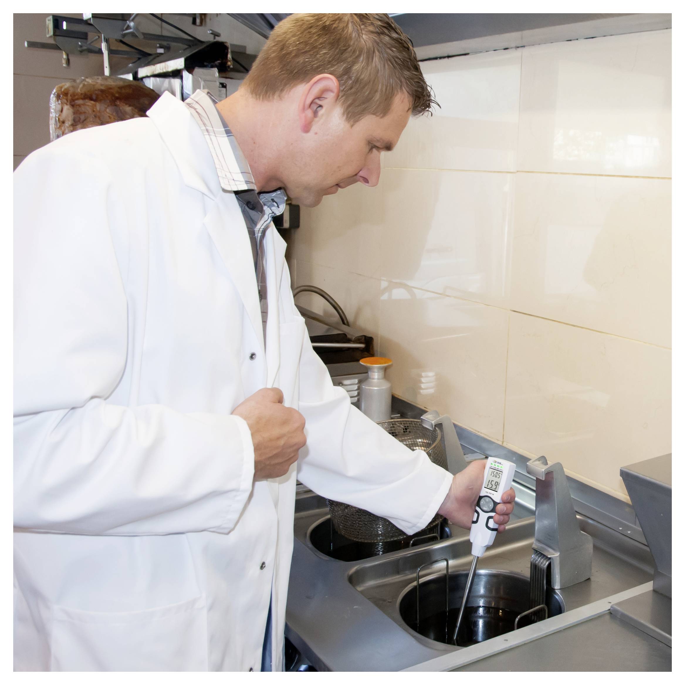 Man in a white lab coat uses a handheld thermometer to measure liquid in a kitchen pot on a stovetop.
