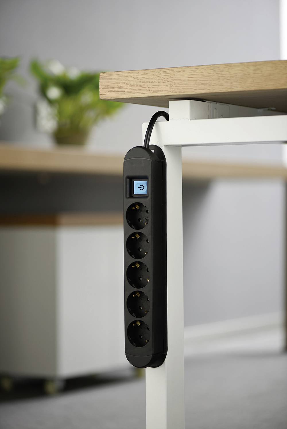 Black power strip with five sockets and a switch, mounted on a white table. Plants and a grey wall in the background.
