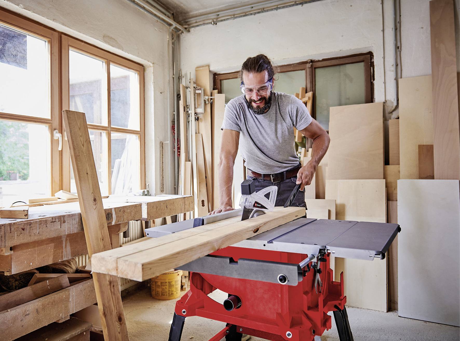 A man is working at a circular saw in a woodwork workshop. He is cutting a long wooden board. The workshop has large windows.