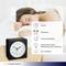 A woman is sleeping next to an analogue alarm clock. The alarm clock has functions such as alarm time, snooze, and light sensor.