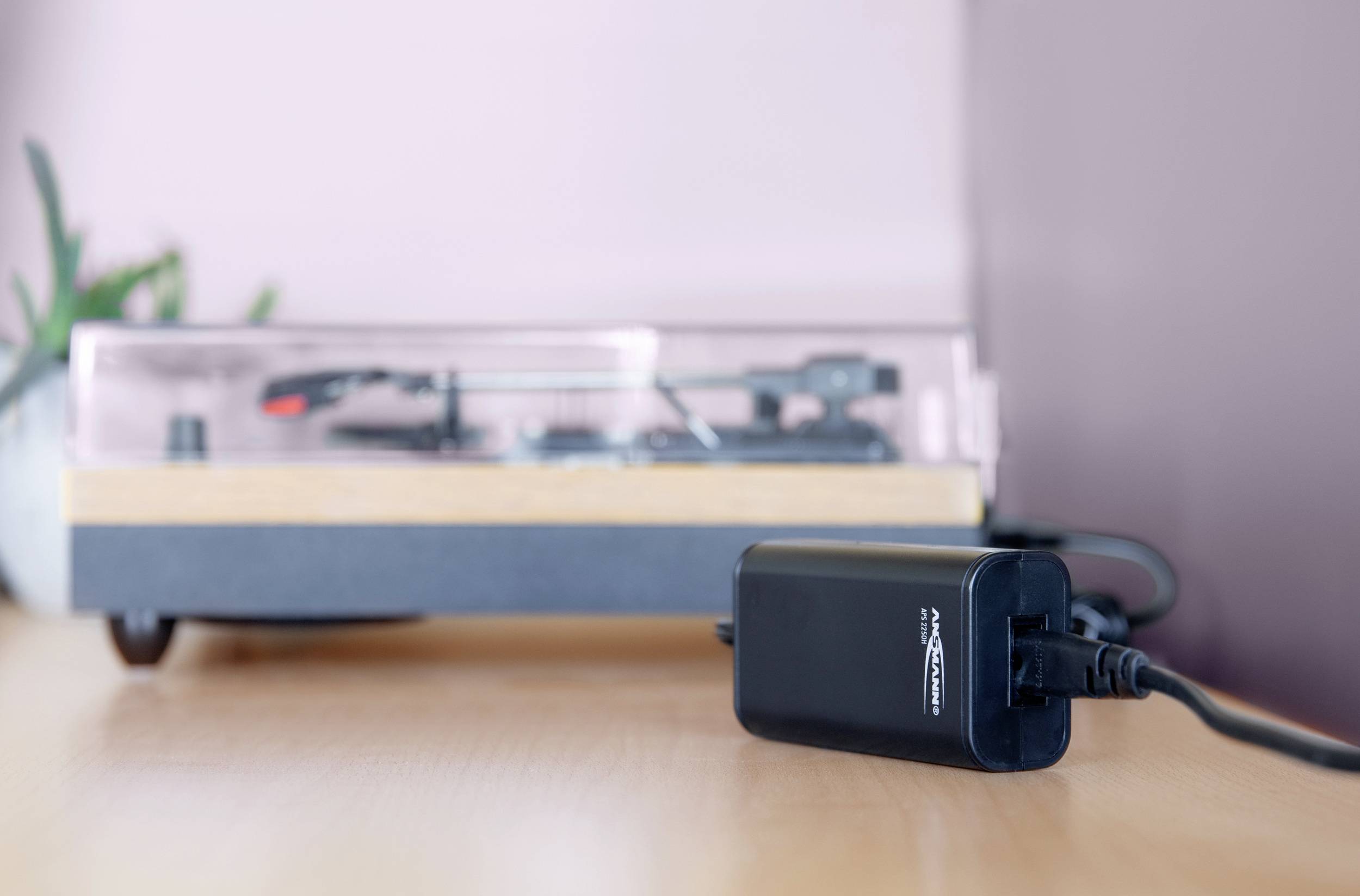 A record player on a table with a connected power supply in the foreground. A houseplant is in the background.