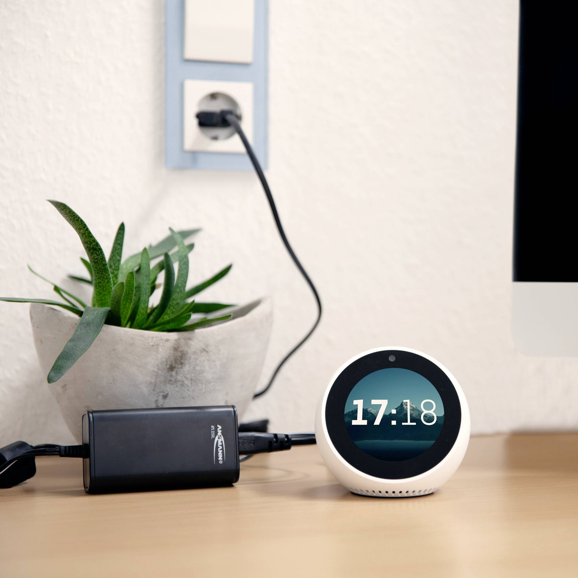 A round digital alarm clock displays the time as 17:18. Next to it sits a plant in a pot, and a charger is plugged in.