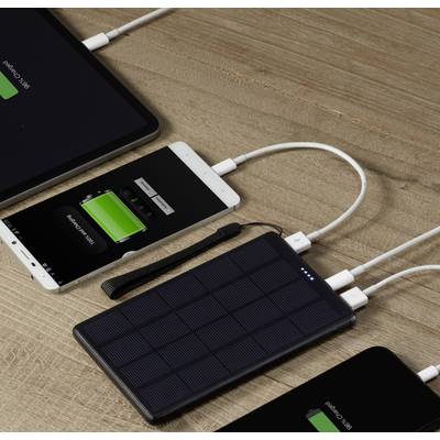 Tablets and smartphones are being charged on a wooden table with a portable solar charger. All devices are showing charging indicators.