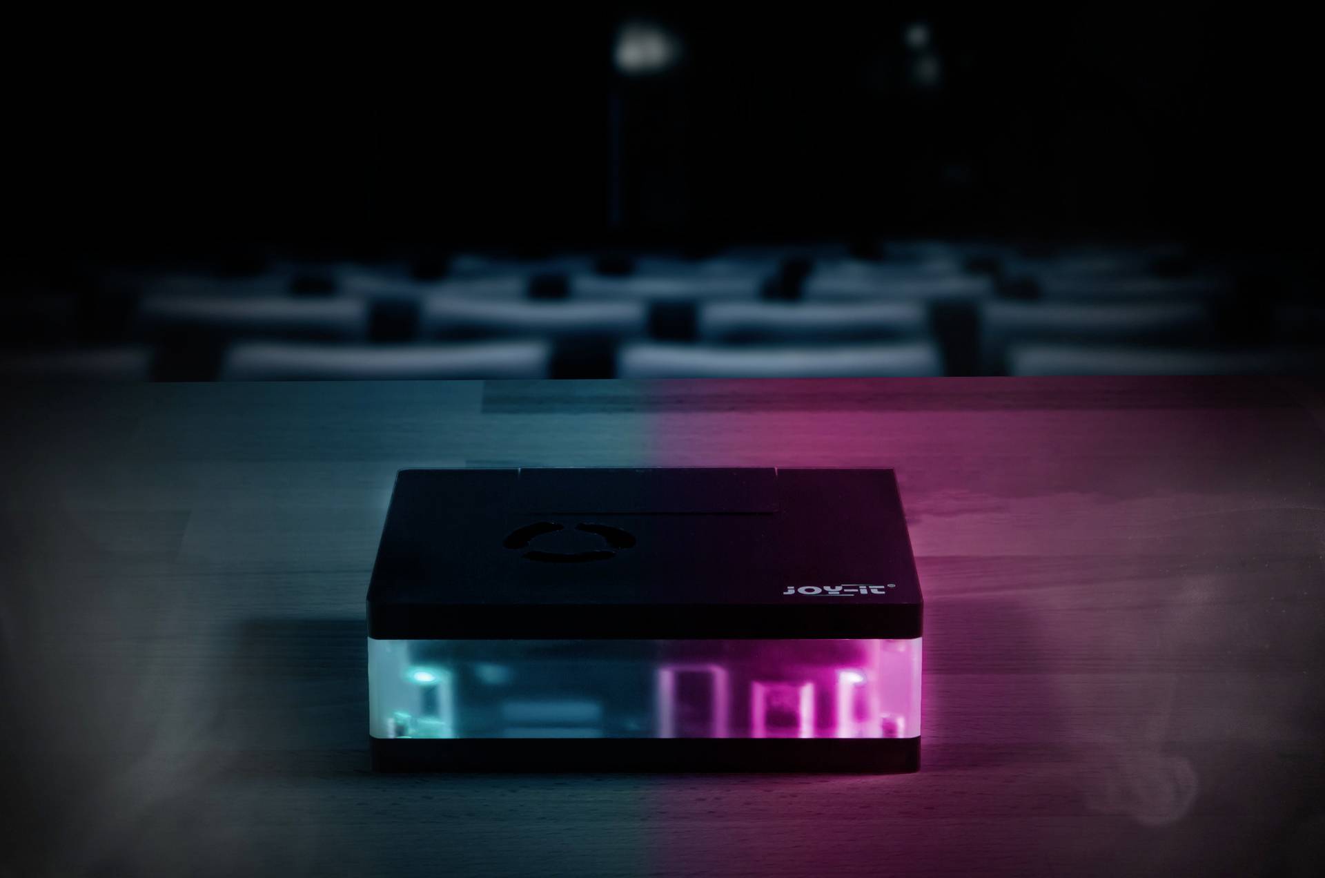 A small, rectangular electronic device with glowing blue-pink LEDs sits on a table in a dark room.