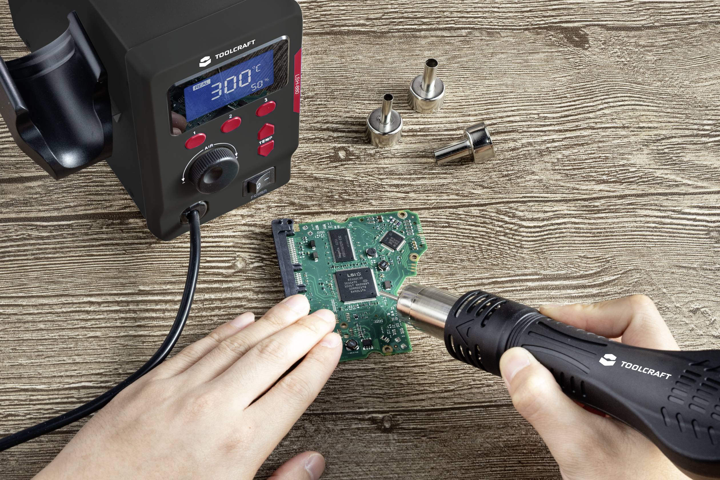 A person is soldering with a hot air gun on a circuit board. A soldering iron and nozzle attachments are lying next to it on the table.