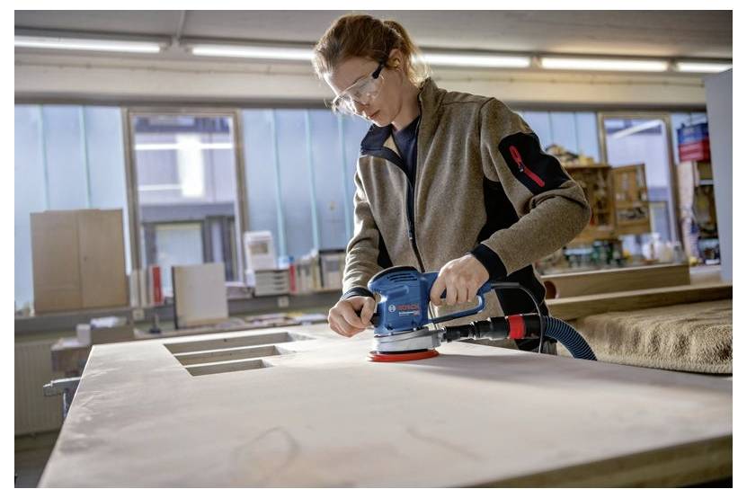 A person wearing safety glasses uses an electric sander on a large wooden board in a workshop setting.