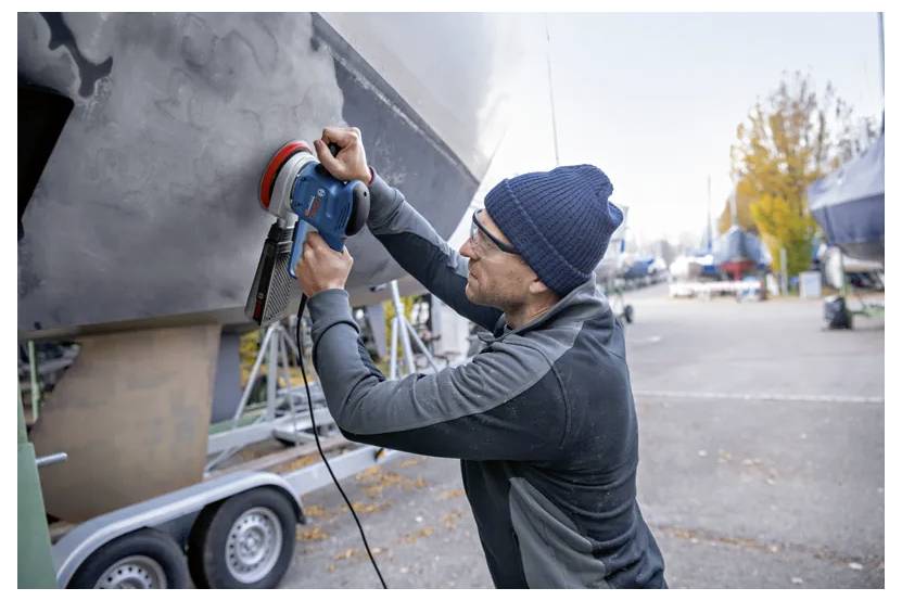 A person wearing a beanie uses a power tool to polish or sand the hull of a boat in a yard with several boats on trailers.