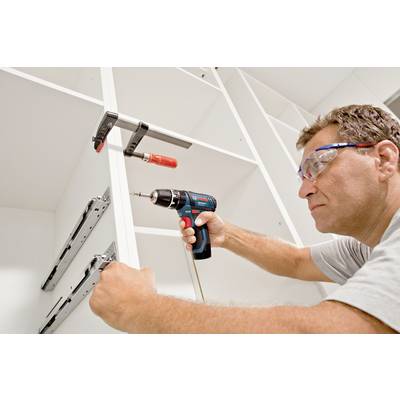 A man is assembling a wardrobe, holding a power drill and wearing safety glasses.