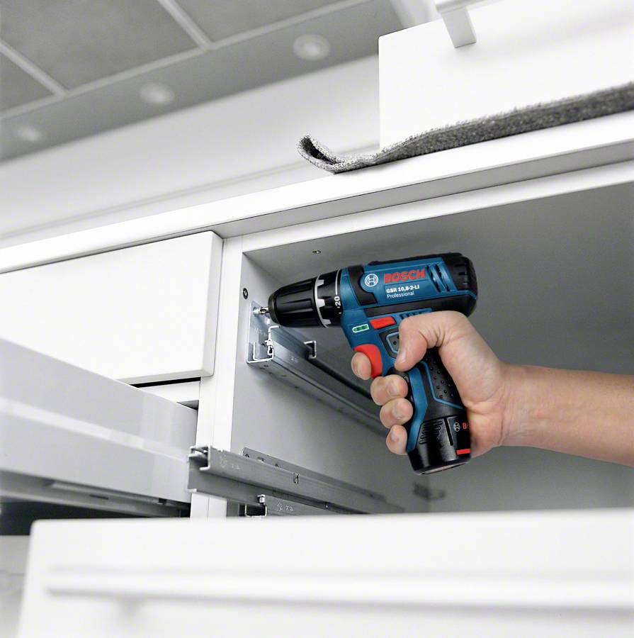 A person is holding a blue cordless drill and attaching a hinge to a white kitchen cabinet door.