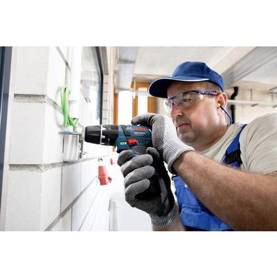 A worker in blue dungarees and safety glasses is drilling into a white brick wall with a cordless drill, holding a green belt.
