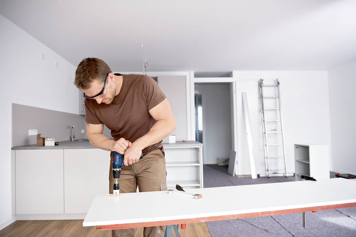 A man is drilling into a white wooden board with a drill in a bright room. Renovation work is in progress in the background.