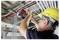 Construction worker in a yellow hard hat uses a power tool to install overhead electrical conduit in a ceiling framework.