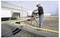 Construction worker drilling conduit through a rooftop pipe bundle beside an industrial building.<br>