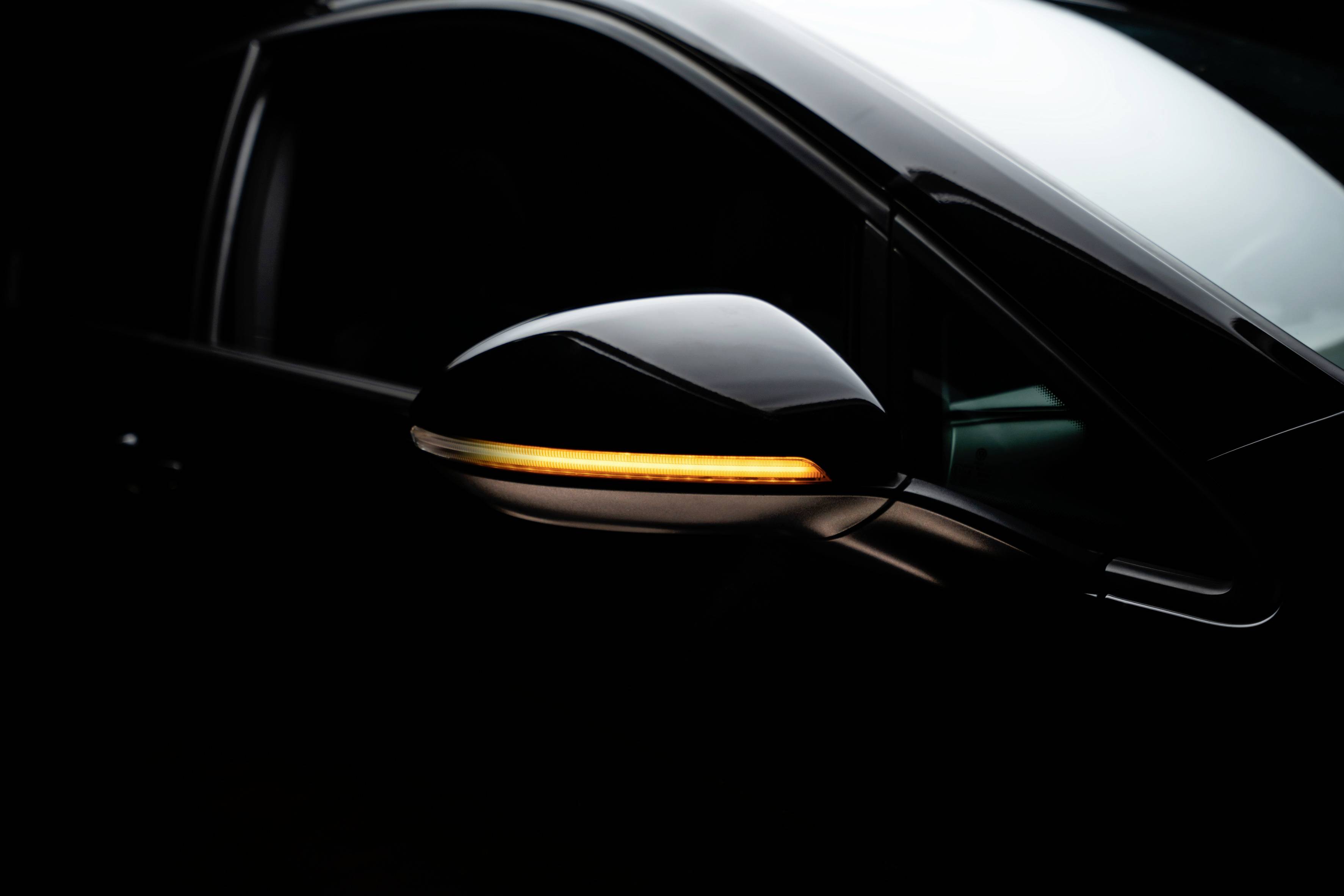 A side wing mirror of a black car shows a bright, orange LED indicator light. Dark background.