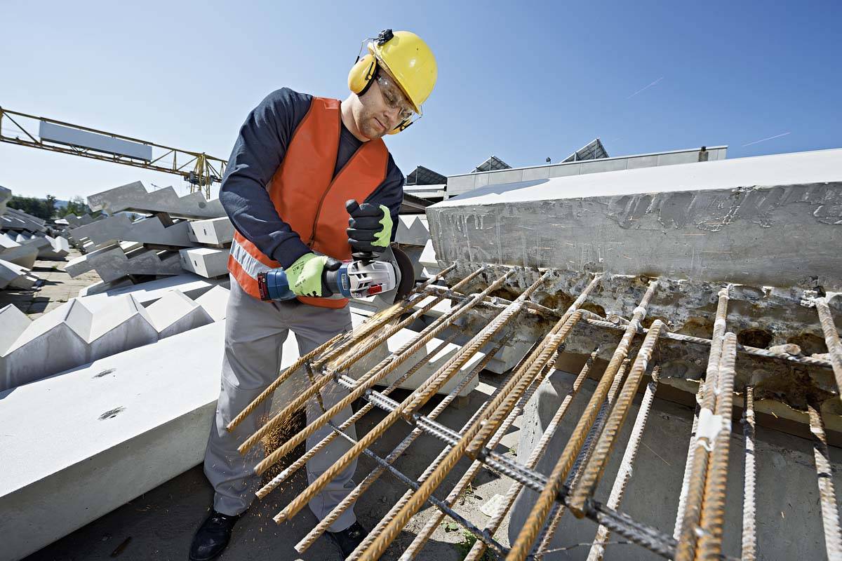 A construction worker in protective clothing cuts rusty reinforcement bars with an angle grinder. Background: concrete slabs on a construction site.
