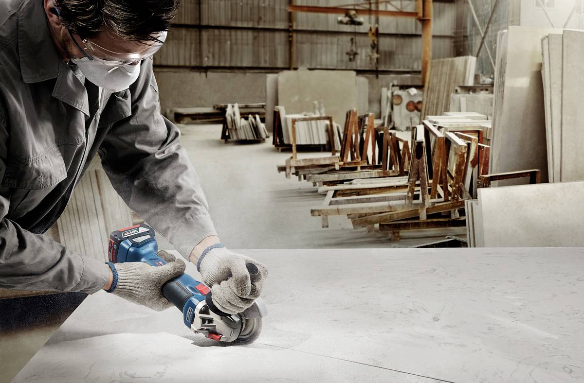 A worker in protective clothing cuts a large stone slab with an electric cutting saw in a factory hall, surrounded by standing materials.