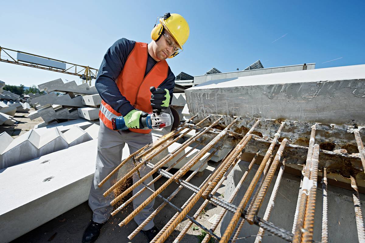 A construction worker wearing a hard hat and high-visibility vest is cutting steel reinforcements with an angle grinder on a building site. Precast concrete elements are lying in the background.