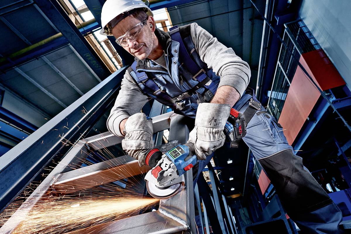 A worker wearing a hard hat and safety glasses is grinding a metal handrail with an angle grinder in a factory environment.