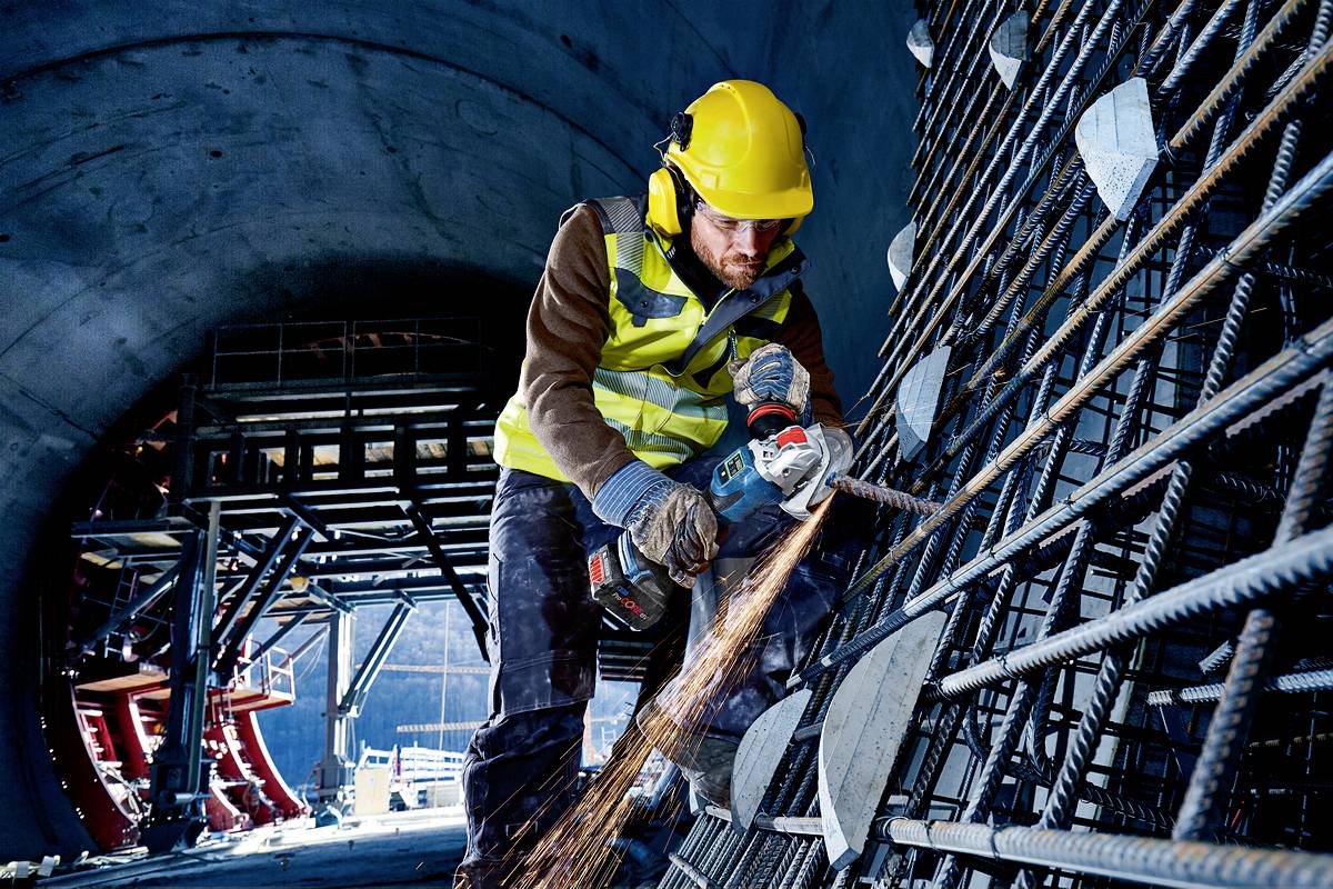 A construction worker cuts metal rods with an angle grinder in a tunnel. Sparks are flying, and he is wearing protective clothing and a hard hat.